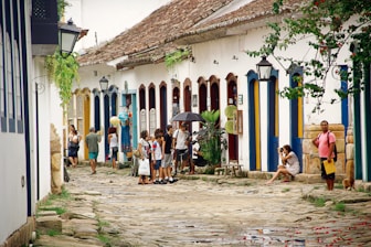 A cobblestone street flanked by colorful colonial buildings with vibrant blue and yellow doors. People are casually walking and socializing, with one person holding an umbrella. Plants and lanterns adorn the street, and a woman is sitting with a camera. The scene conveys a relaxed and historical ambiance.