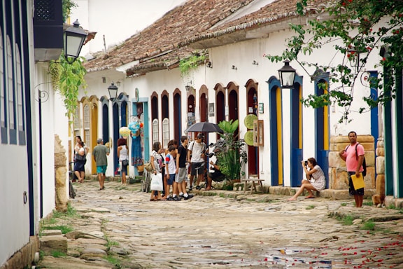 A cobblestone street flanked by colorful colonial buildings with vibrant blue and yellow doors. People are casually walking and socializing, with one person holding an umbrella. Plants and lanterns adorn the street, and a woman is sitting with a camera. The scene conveys a relaxed and historical ambiance.