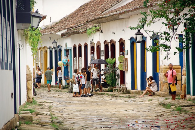 A cobblestone street flanked by colorful colonial buildings with vibrant blue and yellow doors. People are casually walking and socializing, with one person holding an umbrella. Plants and lanterns adorn the street, and a woman is sitting with a camera. The scene conveys a relaxed and historical ambiance.