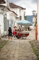 A cobblestone street lined with traditional colonial buildings adorned with colorful shutters and wrought iron lamps. Several people are gathered around a wooden cart with a colorful umbrella, likely selling goods. A woman is walking past, and there is a bicycle leaning against a wall.