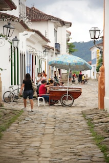 A cobblestone street lined with traditional colonial buildings adorned with colorful shutters and wrought iron lamps. Several people are gathered around a wooden cart with a colorful umbrella, likely selling goods. A woman is walking past, and there is a bicycle leaning against a wall.