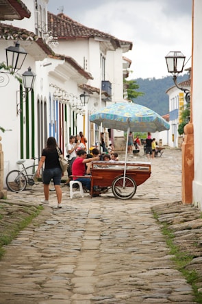 A cobblestone street lined with traditional colonial buildings adorned with colorful shutters and wrought iron lamps. Several people are gathered around a wooden cart with a colorful umbrella, likely selling goods. A woman is walking past, and there is a bicycle leaning against a wall.