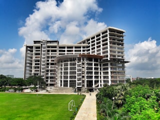 A tall, modern building under construction with scaffolding around several sections. It is surrounded by well-maintained green lawns and trees. The sky is clear with a few fluffy clouds, indicating a sunny day.