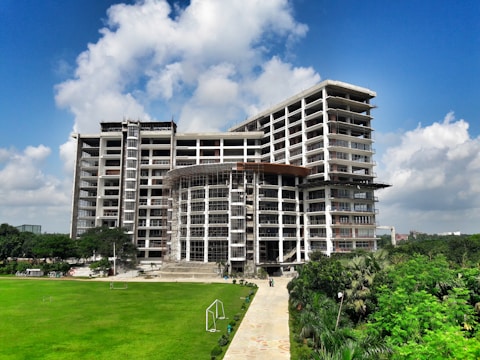 A tall, modern building under construction with scaffolding around several sections. It is surrounded by well-maintained green lawns and trees. The sky is clear with a few fluffy clouds, indicating a sunny day.