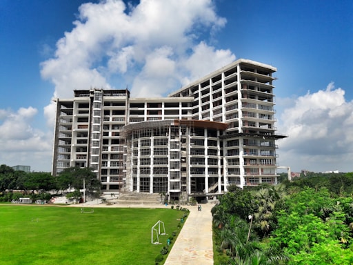 A tall, modern building under construction with scaffolding around several sections. It is surrounded by well-maintained green lawns and trees. The sky is clear with a few fluffy clouds, indicating a sunny day.