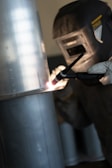 A person is engaged in welding, wearing a protective helmet and gloves. The focus is on the bright arc created during the welding process on a metallic surface.