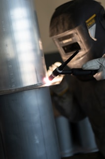A person is engaged in welding, wearing a protective helmet and gloves. The focus is on the bright arc created during the welding process on a metallic surface.