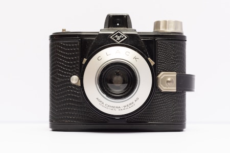 A vintage camera with a black textured body and metallic elements, featuring a prominent lens in the center. The camera has a strap on the right side and a classic design.