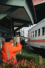 A person wearing a bright orange uniform and a cap stands near a railway track, appearing to be a railway worker or official. They are gesturing with their arm extended as a train passes by. The background includes a small building structure with the sign 'JPL 194' and some greenery.