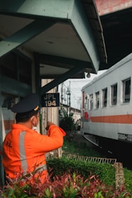 A person wearing a bright orange uniform and a cap stands near a railway track, appearing to be a railway worker or official. They are gesturing with their arm extended as a train passes by. The background includes a small building structure with the sign 'JPL 194' and some greenery.