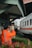 A person wearing a bright orange uniform and a cap stands near a railway track, appearing to be a railway worker or official. They are gesturing with their arm extended as a train passes by. The background includes a small building structure with the sign 'JPL 194' and some greenery.