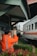 A person wearing a bright orange uniform and a cap stands near a railway track, appearing to be a railway worker or official. They are gesturing with their arm extended as a train passes by. The background includes a small building structure with the sign 'JPL 194' and some greenery.