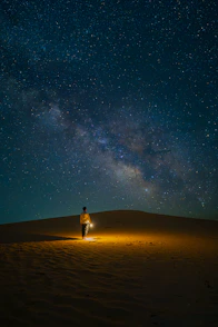 a man standing on top of a sandy hill under a night sky filled with stars