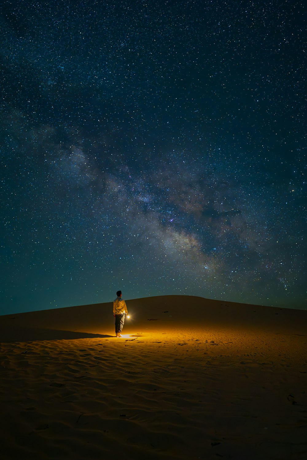 Un homme debout au sommet d’une colline de sable sous un ciel nocturne rempli d’étoiles