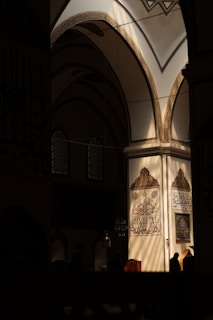 Intricate Arabic calligraphy adorns the walls of a dimly lit mosque interior. Sunlight streams through the arched windows, casting patterns and illuminating the detailed architecture. The atmosphere is serene and contemplative.