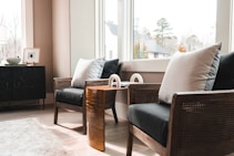 A cozy indoor seating area features two cushioned wooden chairs with neutral colored pillows placed near a large, bright window. A small, stylish side table with a gold finish is situated between the chairs. In the background, a dark wooden console holds decorative items, including framed art and a small bowl. The room has a modern and minimalistic design with outside views of trees and rooftops.