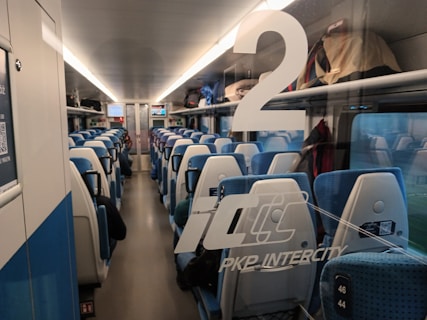 The interior of a modern train carriage features rows of blue upholstered seats with white headrests. Overhead compartments hold various bags and luggage. Train information is visible on digital screens at the far end. The large number '2' is printed on the glass door indicating the carriage class.