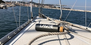 A close-up view of a sailboat's deck with ropes coiled around it. A water bottle labeled 'LAVIE' lies on the deck. The background shows a body of water with a distant shoreline lined with buildings and rocky terrain under a clear blue sky.