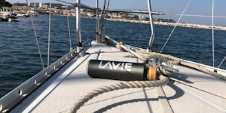A close-up view of a sailboat's deck with ropes coiled around it. A water bottle labeled 'LAVIE' lies on the deck. The background shows a body of water with a distant shoreline lined with buildings and rocky terrain under a clear blue sky.