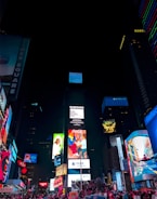 A vibrant urban scene featuring towering skyscrapers adorned with large, colorful digital billboards and advertisements. Crowds of people gather in the illuminated area below, suggesting a bustling, lively atmosphere at night. The bright lights contrast sharply against the dark sky, showcasing famous brands and diverse visual messages.