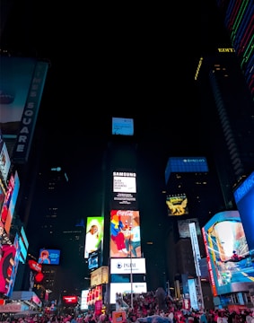A vibrant urban scene featuring towering skyscrapers adorned with large, colorful digital billboards and advertisements. Crowds of people gather in the illuminated area below, suggesting a bustling, lively atmosphere at night. The bright lights contrast sharply against the dark sky, showcasing famous brands and diverse visual messages.