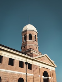 A historic building features a prominent brick tower with a dome-shaped top and arched windows. The architecture showcases classical elements, including columns and decorative stonework. The structure is well-illuminated against a clear blue sky, enhancing its grandeur.