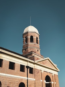 A historic building features a prominent brick tower with a dome-shaped top and arched windows. The architecture showcases classical elements, including columns and decorative stonework. The structure is well-illuminated against a clear blue sky, enhancing its grandeur.