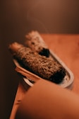 Two bundles of dried herbs are placed on a small, elongated tray. The focus is on the textured surface of the herbs, possibly sage or another smudging herb. A thin wisp of smoke rises gently from one end, indicating recent use. The warm, muted colors provide a calm and soothing atmosphere.