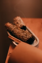 Two bundles of dried herbs are placed on a small, elongated tray. The focus is on the textured surface of the herbs, possibly sage or another smudging herb. A thin wisp of smoke rises gently from one end, indicating recent use. The warm, muted colors provide a calm and soothing atmosphere.