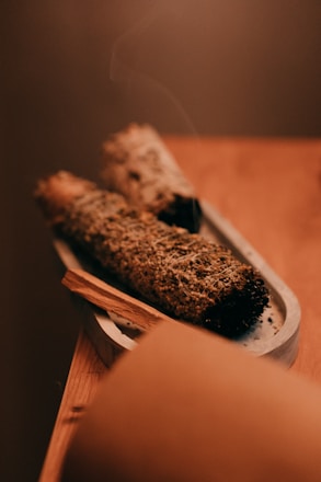 Two bundles of dried herbs are placed on a small, elongated tray. The focus is on the textured surface of the herbs, possibly sage or another smudging herb. A thin wisp of smoke rises gently from one end, indicating recent use. The warm, muted colors provide a calm and soothing atmosphere.