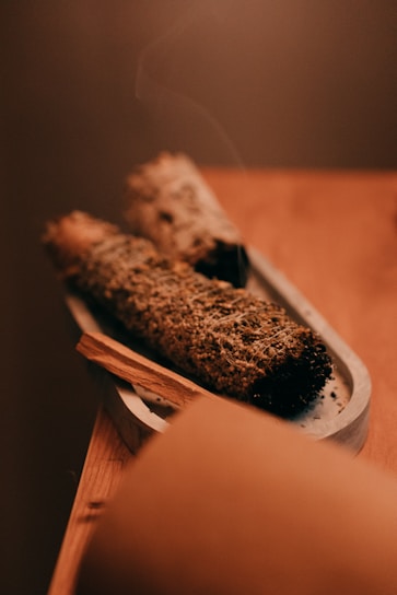 Two bundles of dried herbs are placed on a small, elongated tray. The focus is on the textured surface of the herbs, possibly sage or another smudging herb. A thin wisp of smoke rises gently from one end, indicating recent use. The warm, muted colors provide a calm and soothing atmosphere.