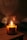 A warmly lit candle labeled 'Tonka + Macadamia' with the words 'Hand Poured Soy Candle' is placed on a wooden surface, emitting a gentle flame. The background is softly blurred, creating a cozy and serene ambiance.