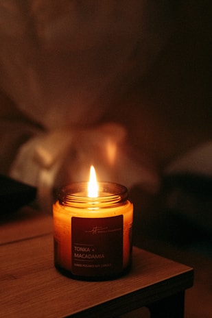 A warmly lit candle labeled 'Tonka + Macadamia' with the words 'Hand Poured Soy Candle' is placed on a wooden surface, emitting a gentle flame. The background is softly blurred, creating a cozy and serene ambiance.