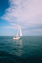 A sailboat with white sails is navigating through calm, blue ocean waters under a partially cloudy sky. The scene is peaceful and expansive, highlighting the open sea and a sense of adventure.