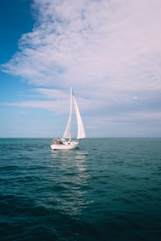 A sailboat with white sails is navigating through calm, blue ocean waters under a partially cloudy sky. The scene is peaceful and expansive, highlighting the open sea and a sense of adventure.
