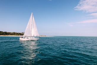 A sailboat gliding past a sunlit Greek island coastline with clear blue waters.
