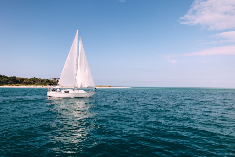 A sailboat gliding past a sunlit Greek island coastline with clear blue waters.