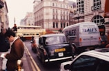 A busy urban street scene with multiple vehicles, including a classic black taxi and a double-decker bus, in a city with historic buildings. A person is standing on the sidewalk holding a map or pamphlet. The area is bustling with traffic and pedestrians.