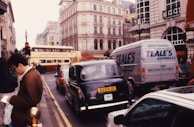 A busy urban street scene with multiple vehicles, including a classic black taxi and a double-decker bus, in a city with historic buildings. A person is standing on the sidewalk holding a map or pamphlet. The area is bustling with traffic and pedestrians.