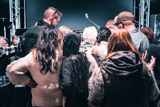 A group of people are gathered closely together in a circle, facing inward. Some have their arms around each other, signifying a sense of unity or support. The background features stage lights and a microphone stand, suggesting they might be at an event or performance.
