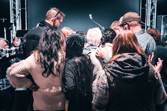 A group of people are gathered closely together in a circle, facing inward. Some have their arms around each other, signifying a sense of unity or support. The background features stage lights and a microphone stand, suggesting they might be at an event or performance.
