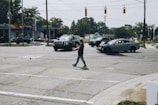 A traffic accident at an intersection involves a damaged gray car with a crumpled front. Police vehicles and officers are present, possibly investigating the scene. A person is walking across the intersection holding a drink. Nearby, a gas station can be seen with trees and residential buildings in the background.