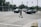 A traffic accident at an intersection involves a damaged gray car with a crumpled front. Police vehicles and officers are present, possibly investigating the scene. A person is walking across the intersection holding a drink. Nearby, a gas station can be seen with trees and residential buildings in the background.