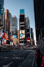A bustling urban scene featuring tall skyscrapers adorned with bright billboards and advertisements. The street is busy with cars and pedestrians. Notable brands and colorful signs dominate the skyline, reflecting a vibrant, city atmosphere.