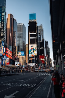 A bustling urban scene featuring tall skyscrapers adorned with bright billboards and advertisements. The street is busy with cars and pedestrians. Notable brands and colorful signs dominate the skyline, reflecting a vibrant, city atmosphere.