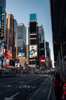A bustling urban scene featuring tall skyscrapers adorned with bright billboards and advertisements. The street is busy with cars and pedestrians. Notable brands and colorful signs dominate the skyline, reflecting a vibrant, city atmosphere.