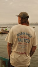 A person with long hair, wearing a cap and a t-shirt, stands by the edge of a body of water. The t-shirt features a design promoting a tattoo parlor with details about its location and opening hours. In the background, several small boats float on the water under a cloudy sky.