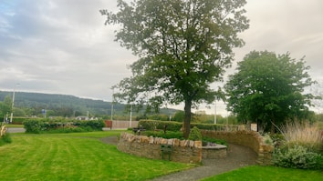 A serene landscape with a large tree standing in the center of a grassy area bordered by a low stone wall. The lush green grass and trees are complemented by a backdrop of rolling hills and a cloudy sky. There is a paved pathway that curves around the tree and leads to other parts of the landscape, creating a peaceful atmosphere.