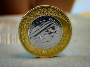 A close-up image of a coin showcasing detailed engravings and Arabic calligraphy. The coin has a dual-tone design with a silver center and a gold outer ring. The face depicted in the center appears to be wearing traditional Middle Eastern attire.