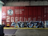 A red freight train car covered with graffiti and the words 'Canadian Pacific Railway' prominently displayed. There are various tags and artistic designs in shades of white and blue on the train car. The setting appears to be inside a warehouse or an industrial area, with a metal roller door and a trash bin visible to the side. The floor consists of concrete with a yellow safety line along the edge.